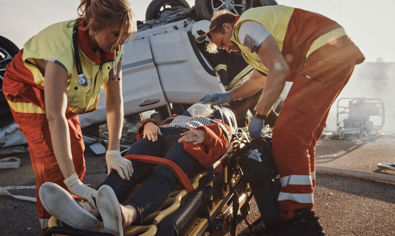On the Car Crash Traffic Accident Scene: Paramedics Saving Life of a Female Victim who is Lying on Stretchers. They Apply Oxygen Mask and Give First Aid. In Background Rollover Vehicle